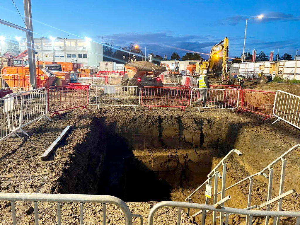 Deep excavation on a building site with a security fence and a dump truck full of soil in the background. DDG Group groundworks.