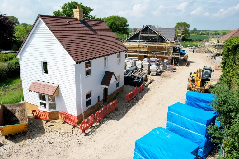 White house on a newly built construction site, surrounded by palettes of materials and a second house in the background with scaffolding up. DDG Group groundworks.