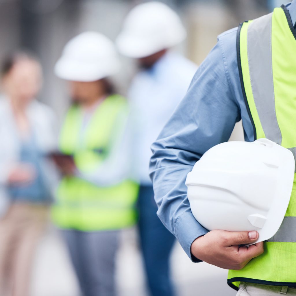 Health and Safety Site expert with white hard hat under their arm and blurred team in the background.